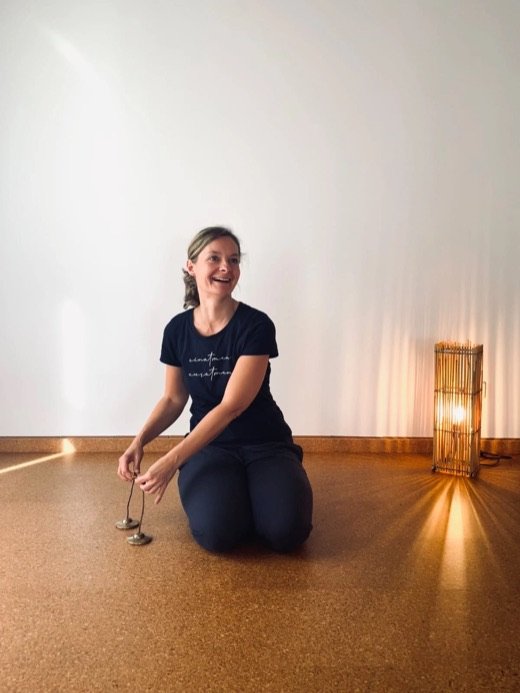 Anja lacht und hält zwei kleine Klangschalen in den Händen, daneben leuchtet eine Bambuslampe